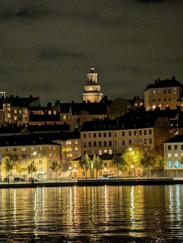 Södermalm at night: the lit tower and dome of Katarina Church above rows of warm-lit buildings and autumn trees on the waterfront, with golden reflections on the dark water.