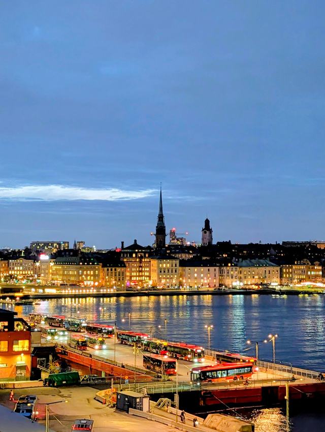 Stockholm at dusk: the Old Town across the water with lit waterfront buildings and a tall church spire under a pale blue sky, red buses waiting on the quay in the foreground.