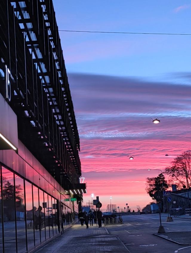 A pavement beside a dark glass building leading towards a sky striped in pink, purple and blue at sunset, with street lamps and people walking in the distance.