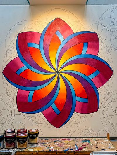 Large painted flower on a wall with eight swirling petals in red, orange, yellow and blue, with jars of paint and brushes on a table below.